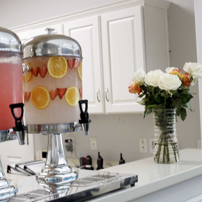 Two drink dispensers with fruit-infused water and a vase of roses on a white countertop in a kitchen.