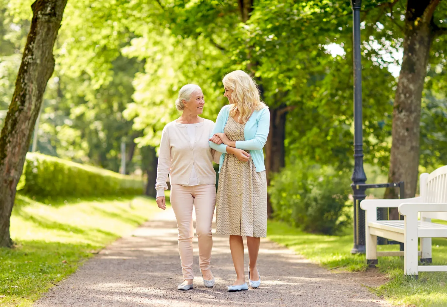 Two women, one older and one younger, walking arm in arm on a sunny park path surrounded by greenery.