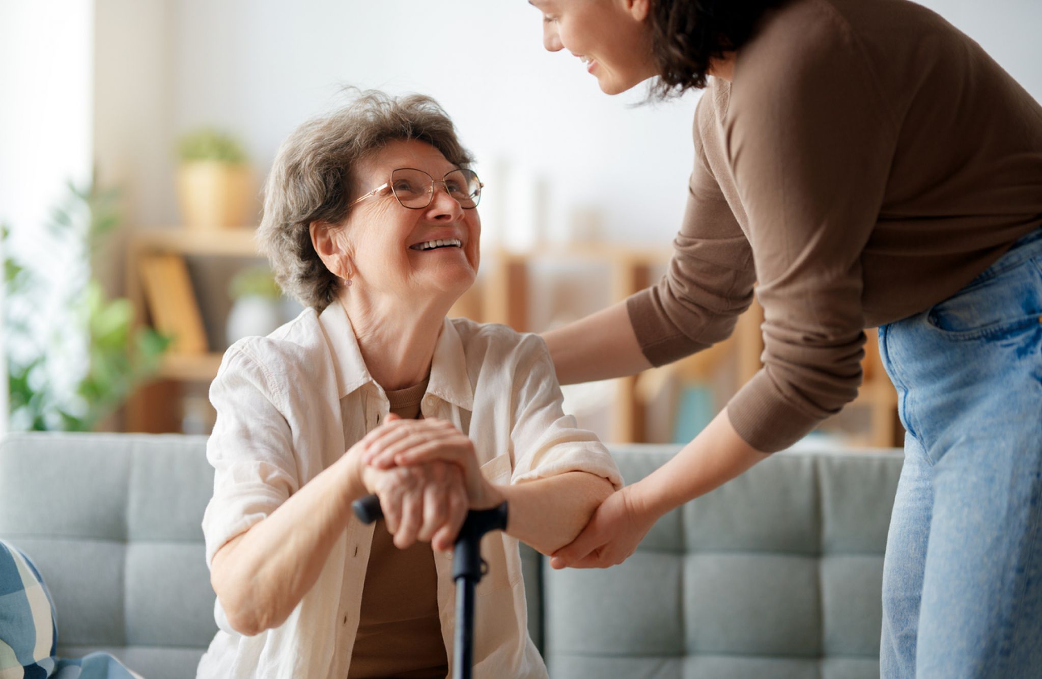 A woman helps her aging mother sit down on the couch, acting as a family caregiver.