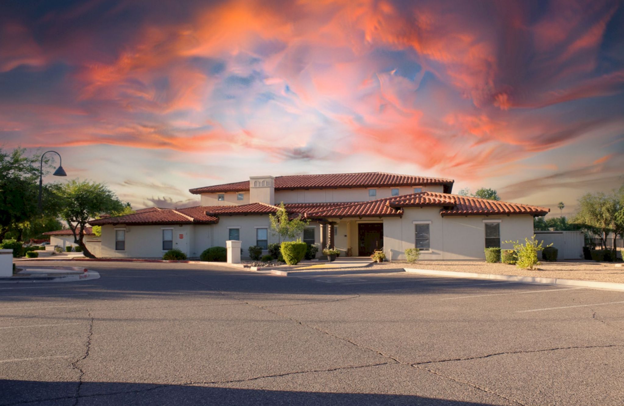 Barton House stands as a Southwestern-style building under a dramatic, colorful sunset sky, with trees and an empty parking lot in front.