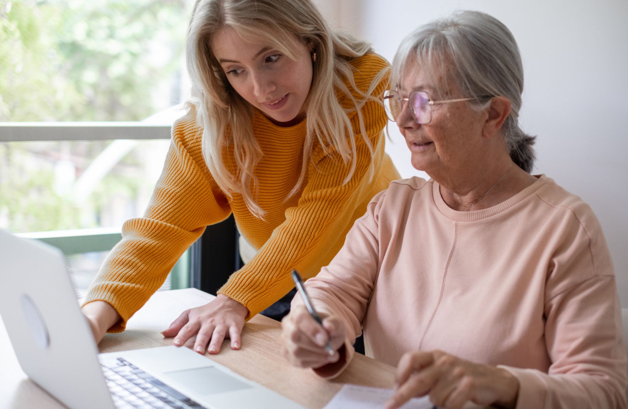 Older woman writes notes while younger woman helps her use a laptop at a table in a bright room.