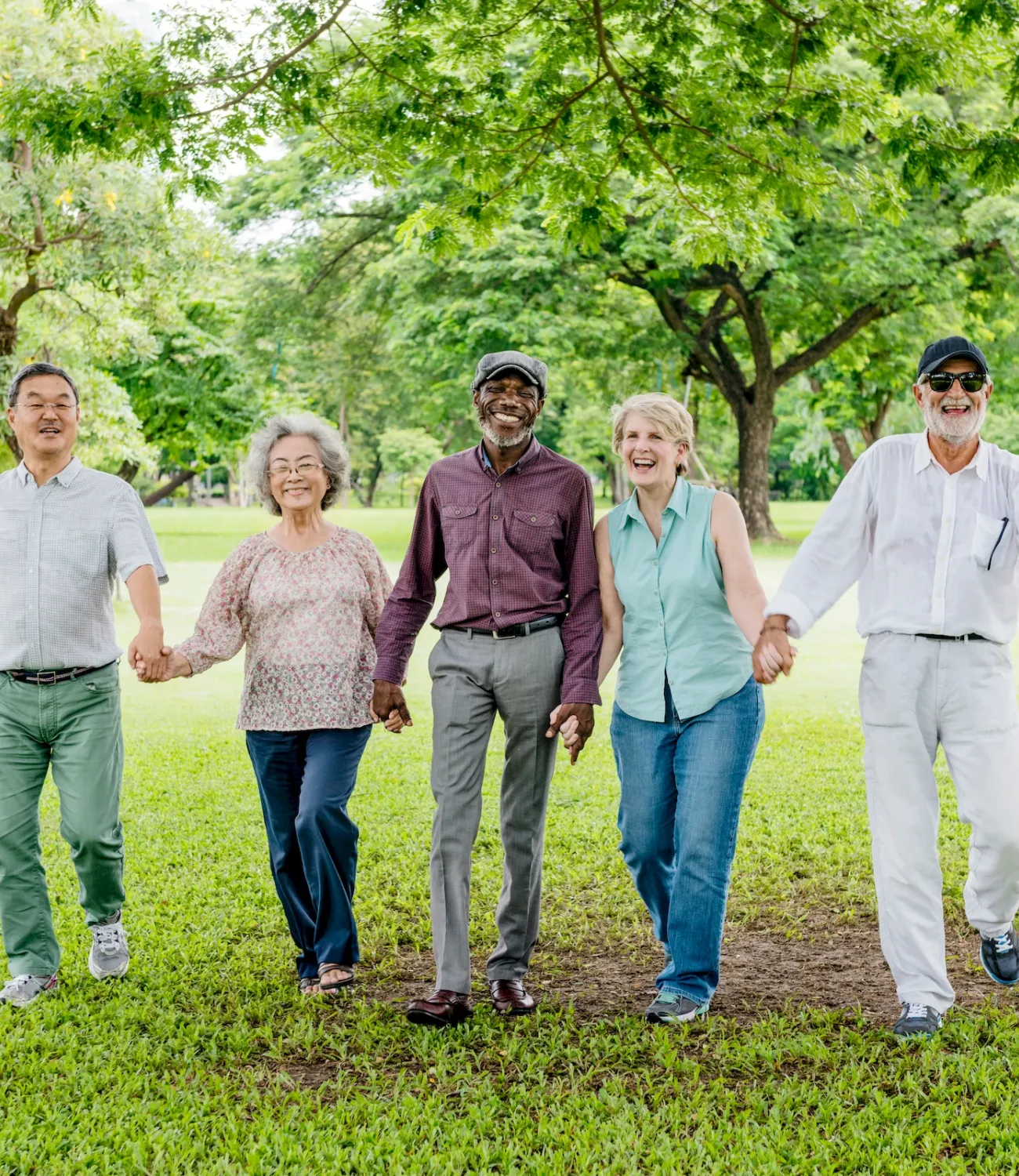 Five smiling adults walk hand in hand through a green park with trees and grass on a sunny day.