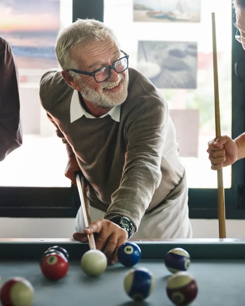 Three older men happily playing pool together in a sunlit room, holding pool cues and smiling.