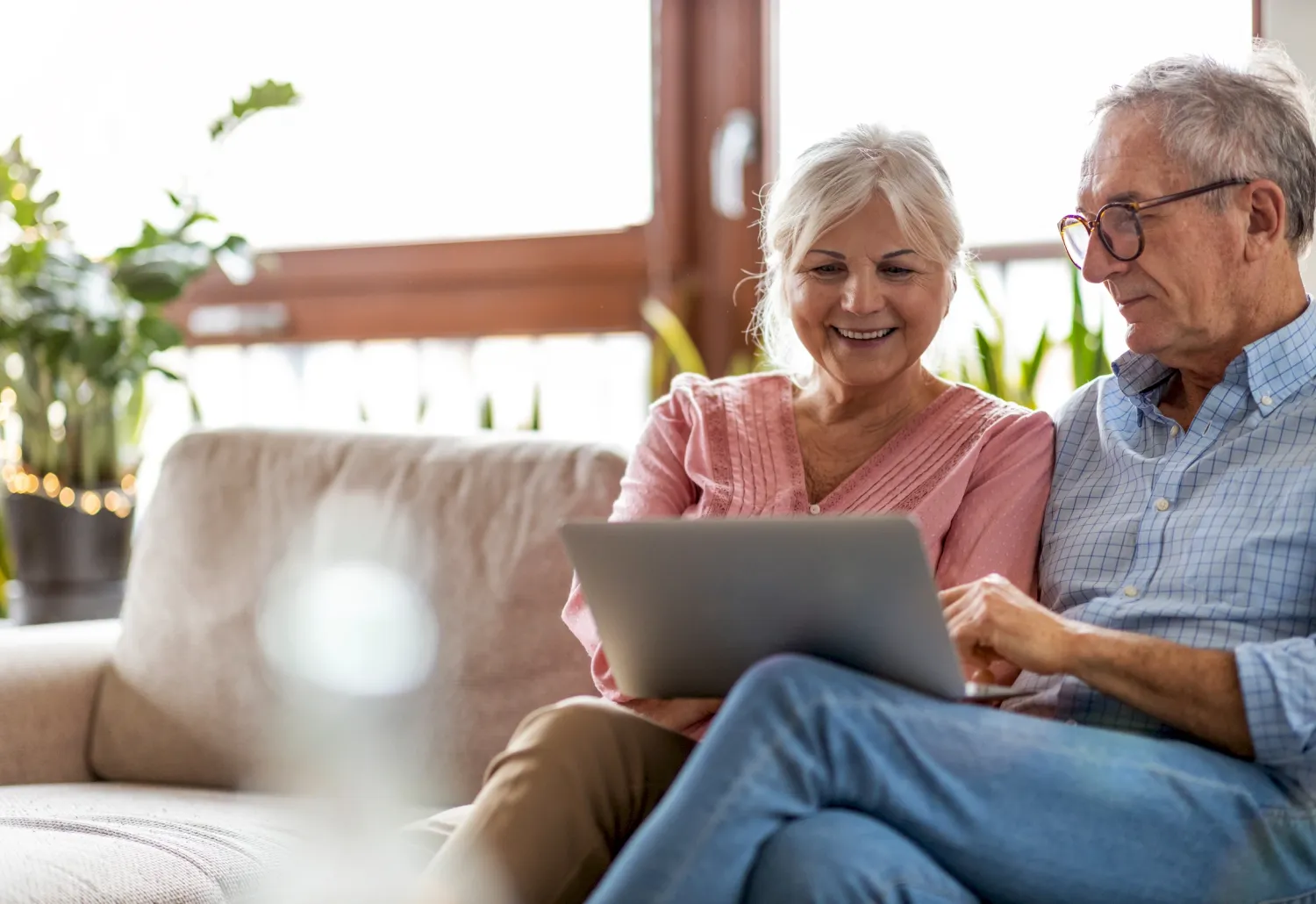 Smiling older couple sitting on a couch, looking at a laptop together in a bright living room.