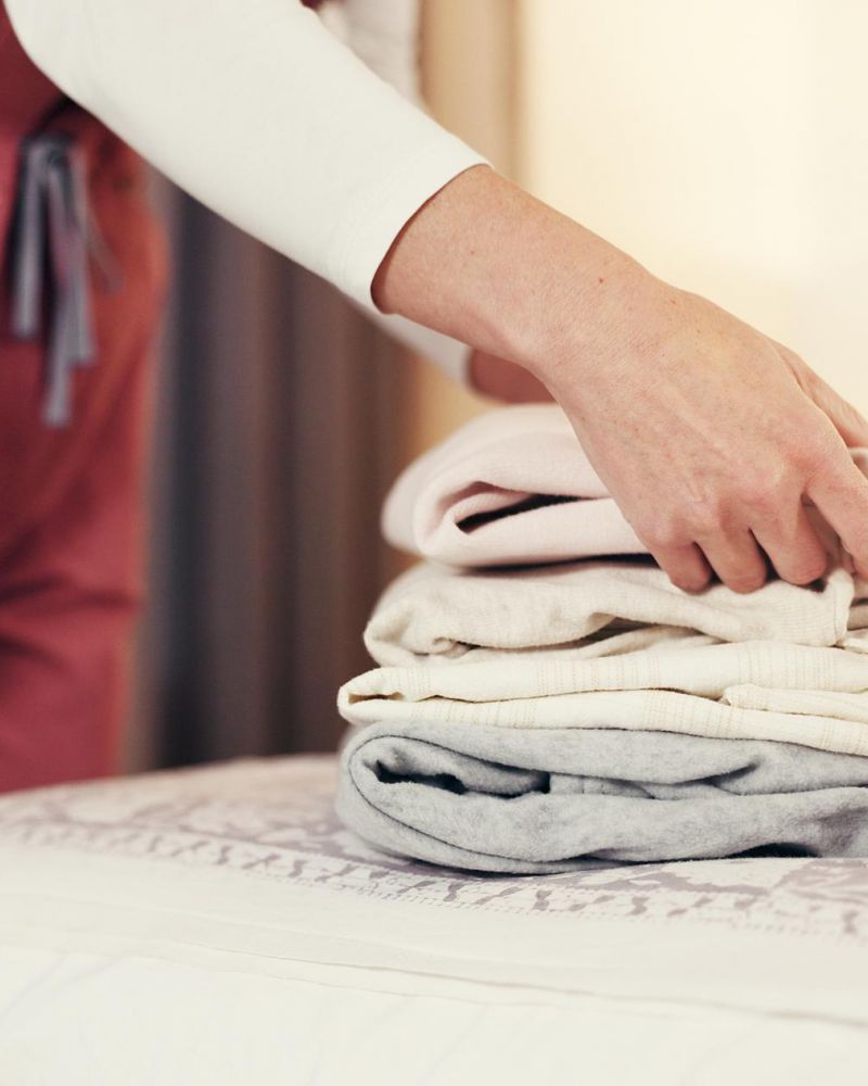 A person folding and stacking clothes on a neatly made bed in a cozy, softly lit room.
