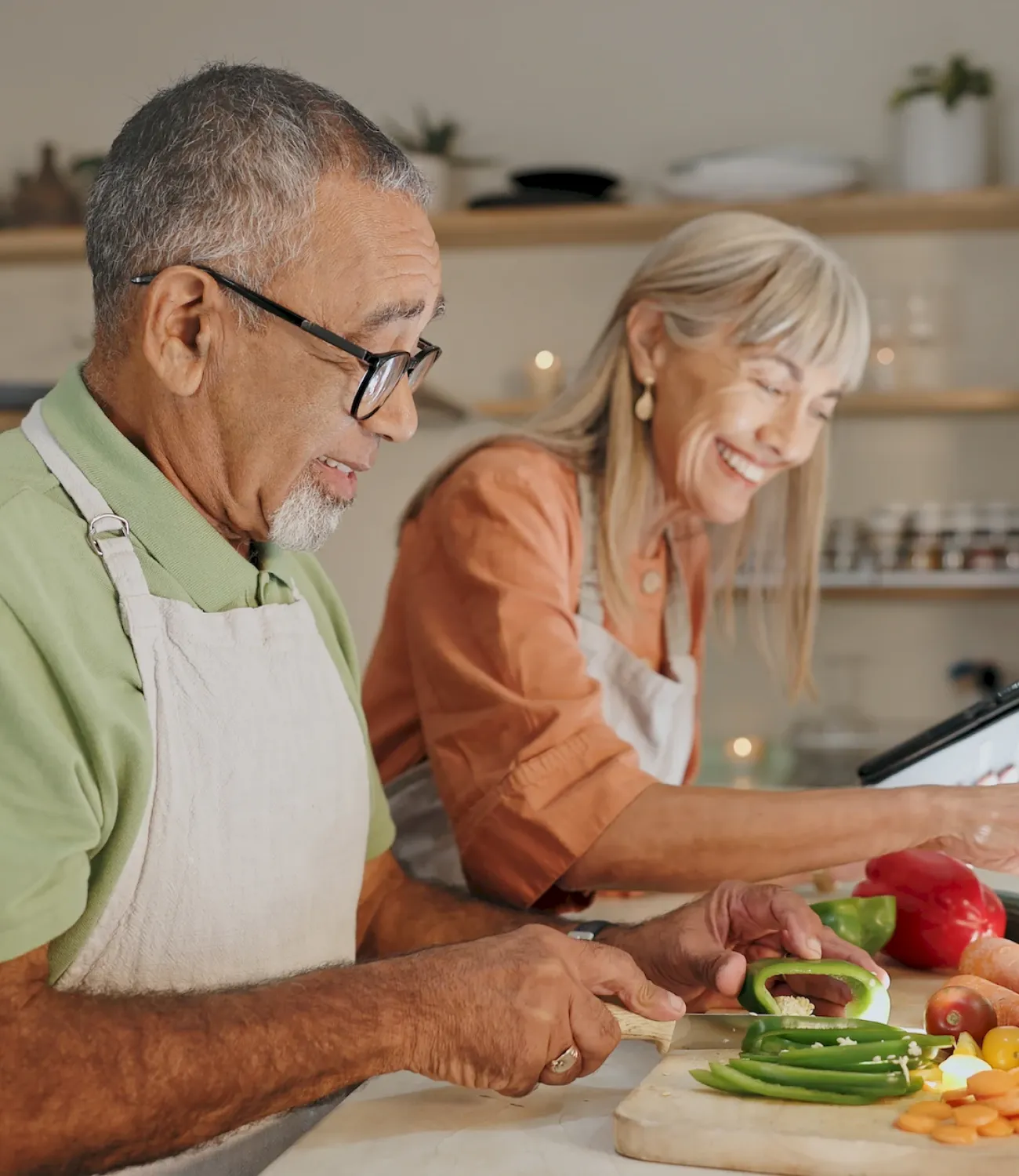Two smiling seniors in aprons chop vegetables together in a bright, modern kitchen.