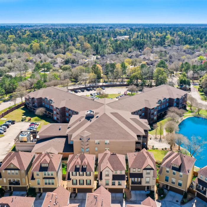 Aerial view of The Terraces at Kingwood, an apartment complex with a pond, surrounded by trees and houses.