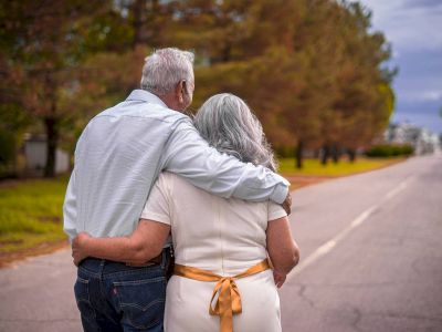 An elderly couple walks arm in arm down a tree-lined road, Honoring Every Story on a cloudy day.
