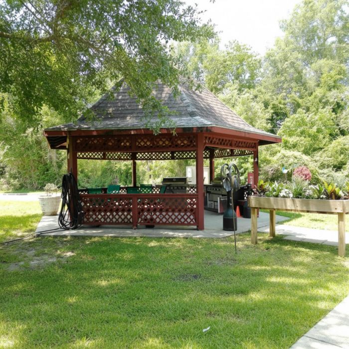 A red gazebo with garden tools and a raised planter, nestled in Tarina of Kingwood, surrounded by grass and trees on a sunny day.