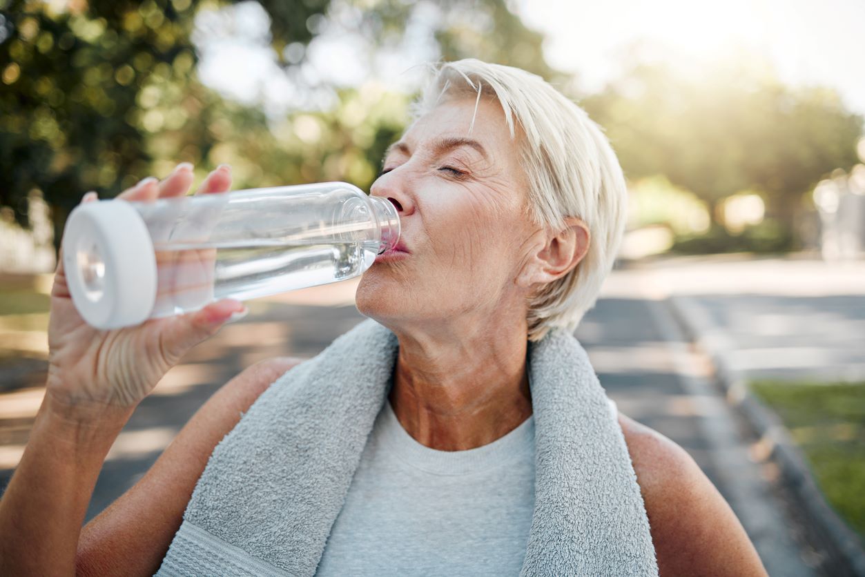 An older woman sips from a glass water bottle to rehydrate while exercising.