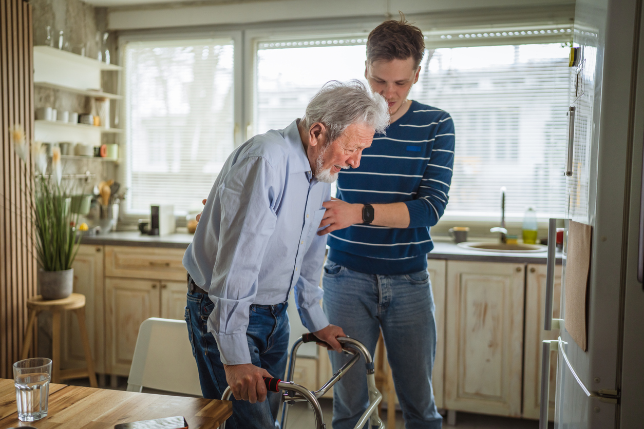 Older adult using a walker while another person provides support indoors. 
