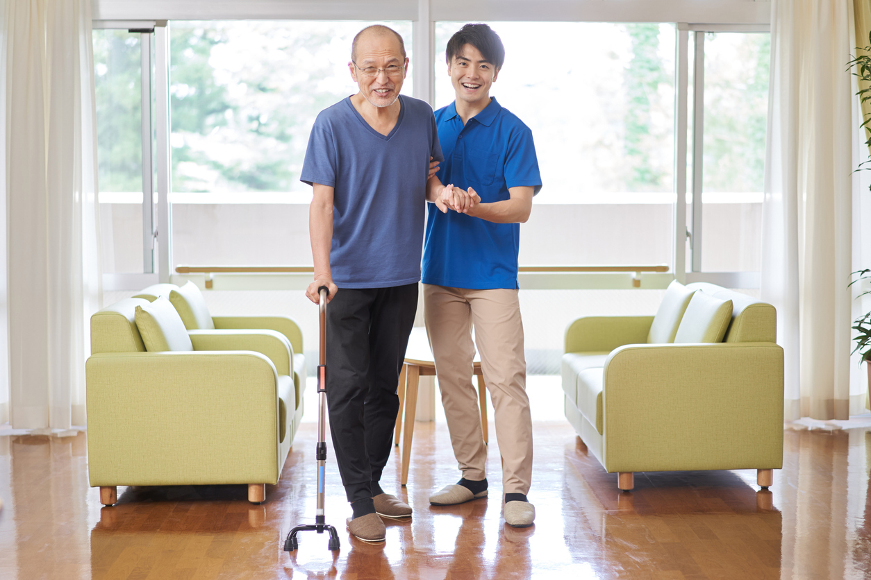 An older man with a cane is assisted by a younger man in an assisted living, modern room with green chairs.