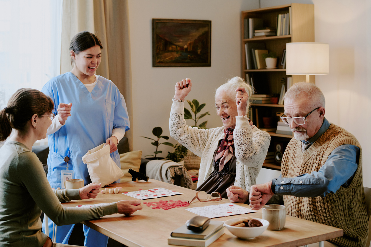 Four seniors play bingo as one cheers, a nurse smiles nearby, highlighting the difference in Assisted Living and Memory Care settings.